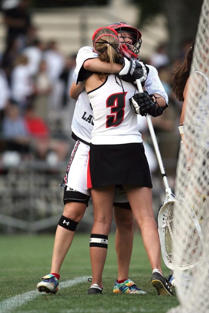 Two female lacrosse players celebrating victory on the field, embracing warmly.